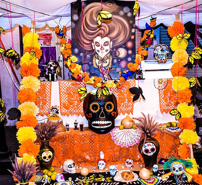 An altar for Dia de los Muertos covered in yellow and orage paper marigold flowers painted ceramic skills, and food.